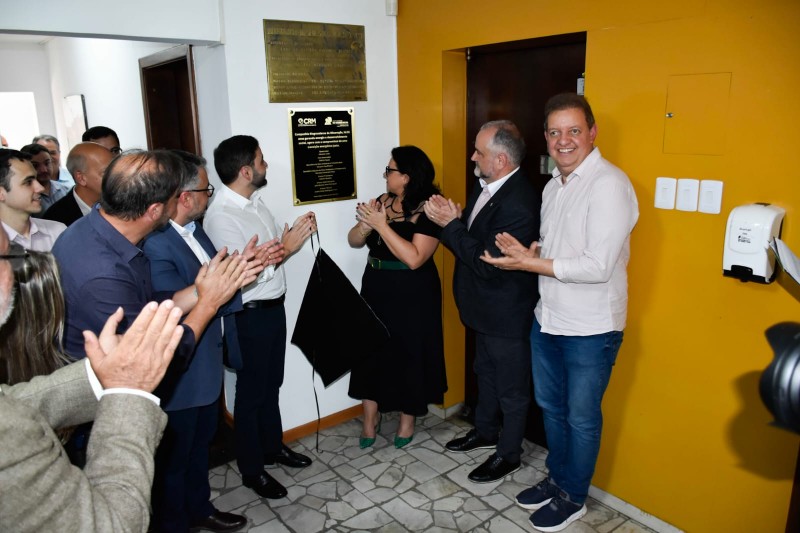 A imagem mostra um grupo de autoridades descerrando uma placa comemorativa dos 55 anos da Companhia Riograndense de Mineração (CRM). Elas posicionadas ao redor da placa, batendo palmas. A placa está em posição vertical em uma parede. onde a placa foi coberta com um tecido, que foi retirada para revelação. Todos estão vestidos informalmente e alguns homens com blazer e camisa social