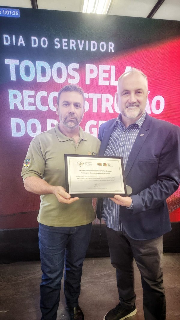 A imagem mostra o Presidente Ademir Baretta ao lado do funcionário Luis Adelcio. Ambos sorrindo e seguram juntos uma placa de homenagem, com os olhares voltados para a câmera. Luis Adelcio, que recebeu essa homenagem após uma votação online, representa a subsecretaria ou departamento reconhecido pelo importante papel desempenhado no enfrentamento dos efeitos do desastre meteorológico causado pelas enchentes. A placa contém detalhes do prêmio e o nome do órgão, destacando o esforço conjunto da equipe. Ambos os homens vestem roupas formais, com a expressão de satisfação.
