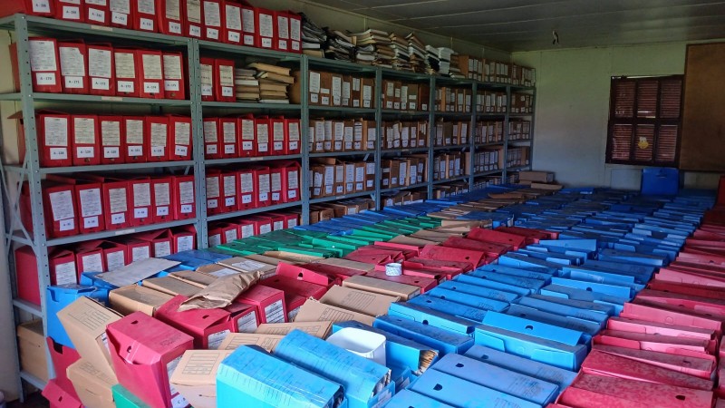 Imagem mostrando vários arquivos organizados no chão e em prateleiras na vertical após uma enchente que danificou grande parte do patrimônio do CRM. Os documentos estão separados em pastas de plástico, tipo caixa em azul, vermelho e pardo, para triagem ou secagem.