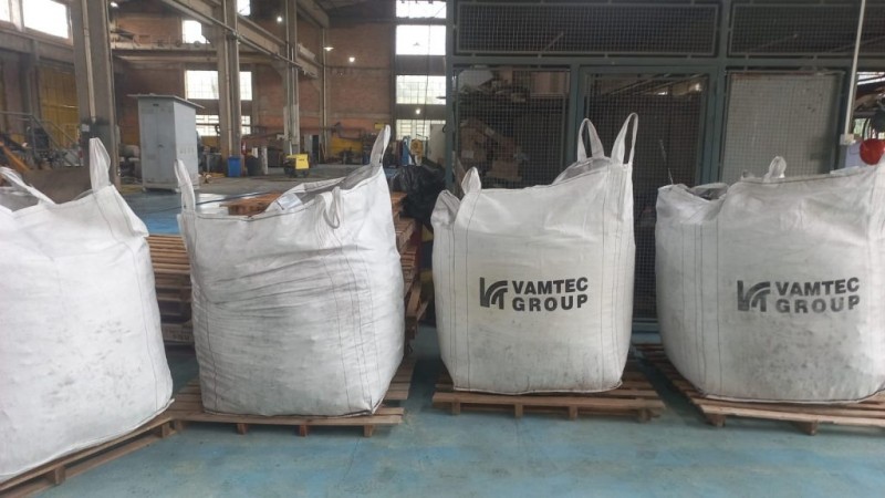 Imagem de várias sacolas lado a lado contendo carvão mineral, prontas para envio. Nas sacolas, está escrito "Vamtec Group" em destaque. O destino mencionado é Karaganda, no Cazaquistão. As sacolas estão organizadas em um ambiente industrial, sugerindo um local de armazenamento ou expedição.