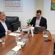 A imagem mostra quatro pessoas reunidas em uma reunião na Procuradoria Geral do Estado (PGE). As pessoas estão sentadas ao redor de uma mesa retangular. À esquerda, há um homem de pele clara, com cabelo curto e usando uma camisa branca. Ao lado dele, uma mulher de pele morena, com cabelo preso em um coque e vestindo uma blusa branca mexendo em papéis. anotações em um bloco. Documentos e canetas estão espalhados sobre a mesa, indicando que a reunião está em andamento.