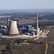Imagem de uma usina termelétrica em funcionamento. No centro, há grandes estruturas industriais com torres metálicas e chaminés altas liberando fumaça branca no céu. À frente, tanques e tubos conectam diferentes partes da planta. Ao fundo, é possível ver linhas de transmissão de energia elétrica e postes conectando a usina à rede. O céu está parcialmente nublado, e o ambiente ao redor é plano, com vegetação esparsa.