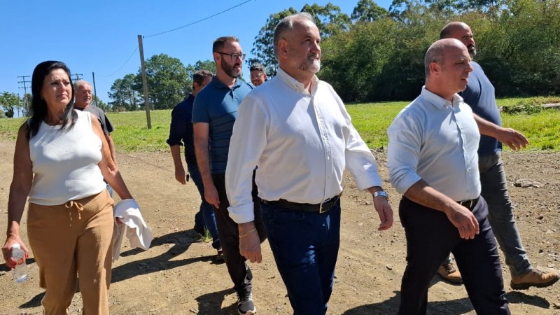 Parte da comitiva de trabalho da Prefeitura de Cachoeira do Sul e gestores da CRM juntos por uma área de mineração. O grupo, composto na maioria por homens e apenas uma mulher estão vestidos com roupas formais, conversam enquanto observam o local. Ao fundo, é possível ver a paisagem típica de revegetação.