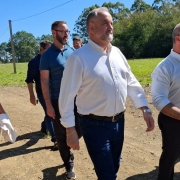 Parte da comitiva de trabalho da Prefeitura de Cachoeira do Sul e gestores da CRM juntos por uma área de mineração. O grupo, composto na maioria por homens e apenas uma mulher estão vestidos com roupas formais, conversam enquanto observam o local. Ao fundo, é possível ver a paisagem típica de revegetação.