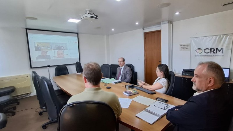 Gestores da CRM e conselheiros de administração participam de uma reunião híbrida. Algumas pessoas estão sentadas ao redor de uma mesa em uma sala de reuniões, enquanto outras aparecem em uma grande tela, conectadas virtualmente. Todos estão atentos, com papéis, notebooks e materiais de apoio sobre a mesa.