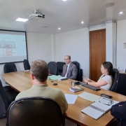 Gestores da CRM e conselheiros de administração participam de uma reunião híbrida. Algumas pessoas estão sentadas ao redor de uma mesa em uma sala de reuniões, enquanto outras aparecem em uma grande tela, conectadas virtualmente. Todos estão atentos, com papéis, notebooks e materiais de apoio sobre a mesa.