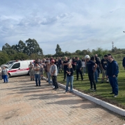 imulado de evacuação de emergência realizado em via pública na cidade de Candiota. A imagem mostra moradores e equipes de resposta caminhando pela rua, seguindo orientações de agentes uniformizados. Algumas pessoas carregam mochilas ou pertences, representando uma evacuação realista. Há viaturas e cones de sinalização no local, e o clima é de organização e atenção. O exercício ocorre durante o dia, com boa visibilidade.