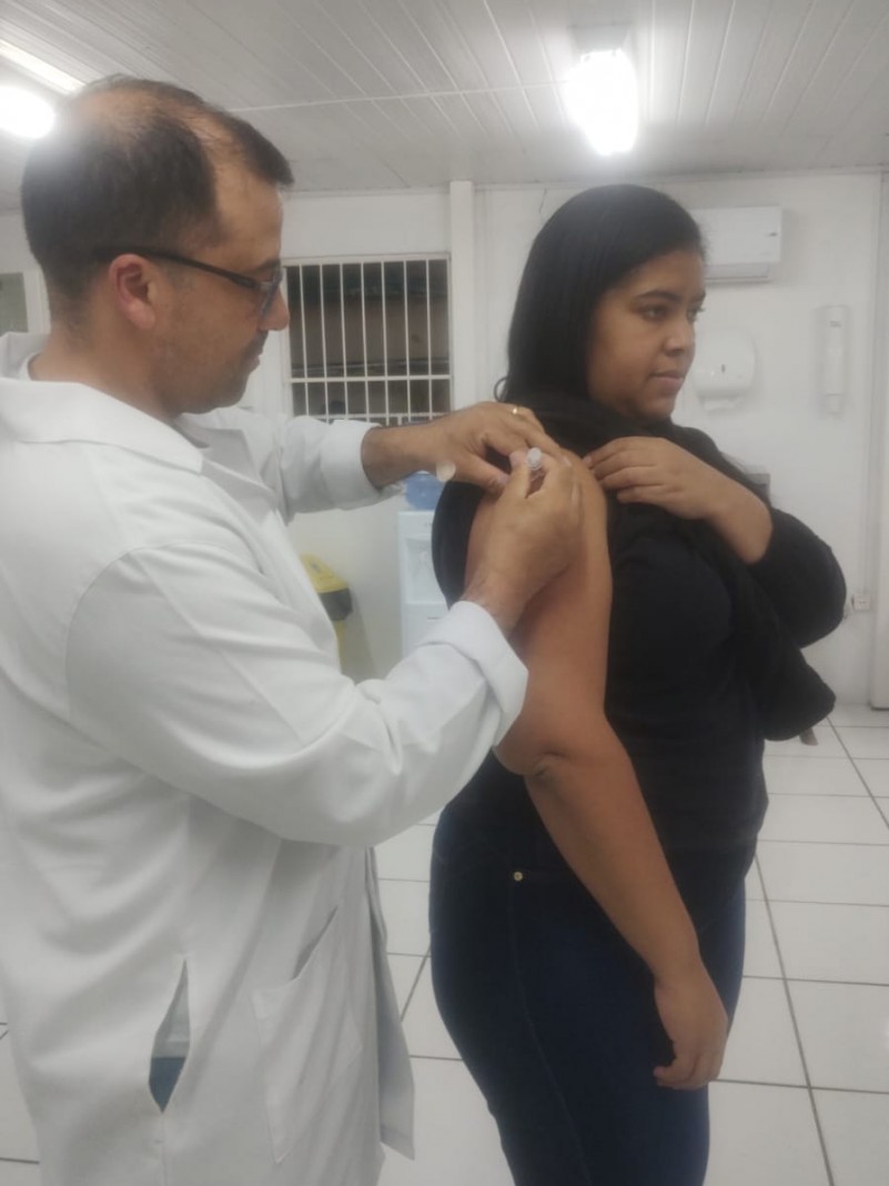 A imagem mostra uma mulher de blusa preta, recebendo a vacina do enfermeiro técnico. Vacina influenza da gripe