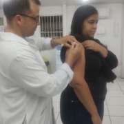 A imagem mostra uma mulher de blusa preta, recebendo a vacina do enfermeiro técnico. Vacina influenza da gripe