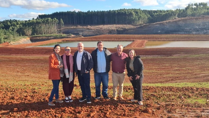 Seis pessoas da equipe da CRM (Companhia Riograndense de Mineração) estão reunidas em uma área de mineração a céu aberto em Candiota. Todos usam roupas normais. Ao fundo, é possível ver as áreas novas mineradas. O grupo está sorrindo e posicionado lado a lado, demonstrando união e profissionalismo no ambiente de trabalho.